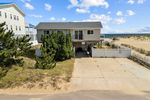 A two-story house with a covered porch, a sandy beach in the background, and a clear sky.