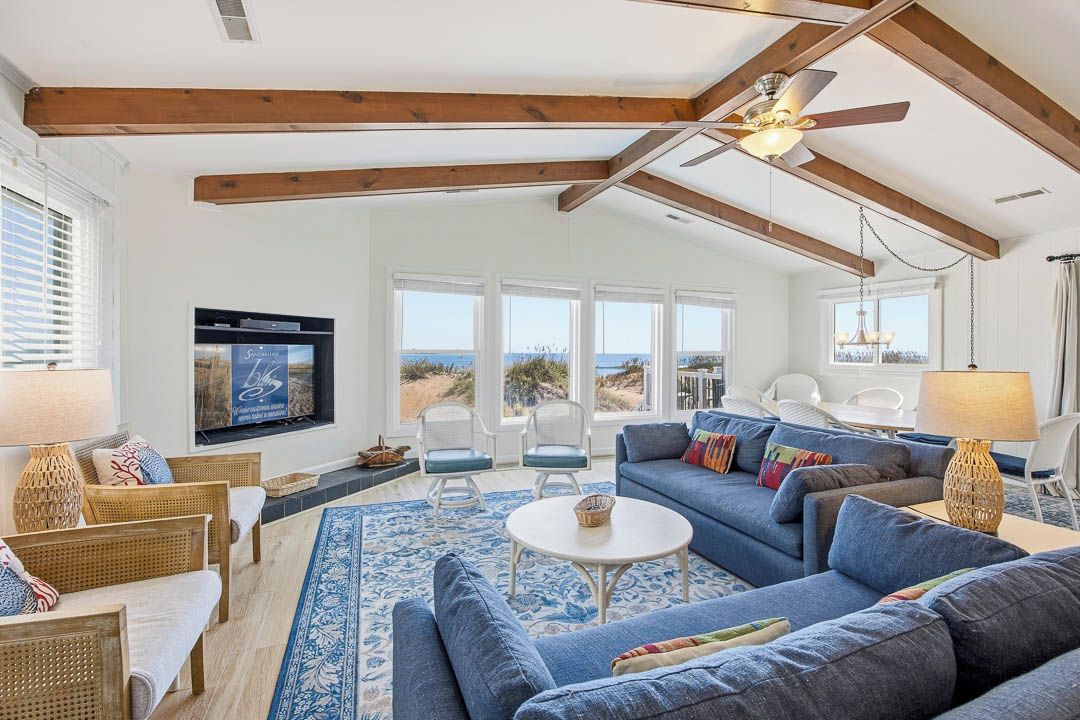 A spacious living room with a large blue sofa, a white coffee table, and wicker furniture. There is a ceiling fan, large windows with a view, and a flat-screen TV mounted on the wall.