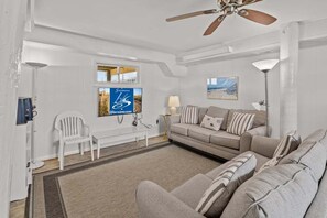 A spacious living room with a sofa, a coffee table, and a white chair. There is a ceiling fan, a window with a view, and a painting on the wall.