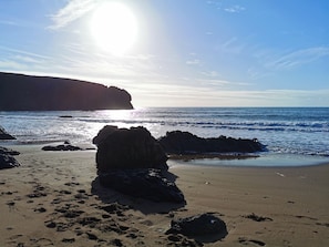 Une plage avec de gros rochers, un ciel dégagé et la lumière du soleil se reflétant sur l’eau.