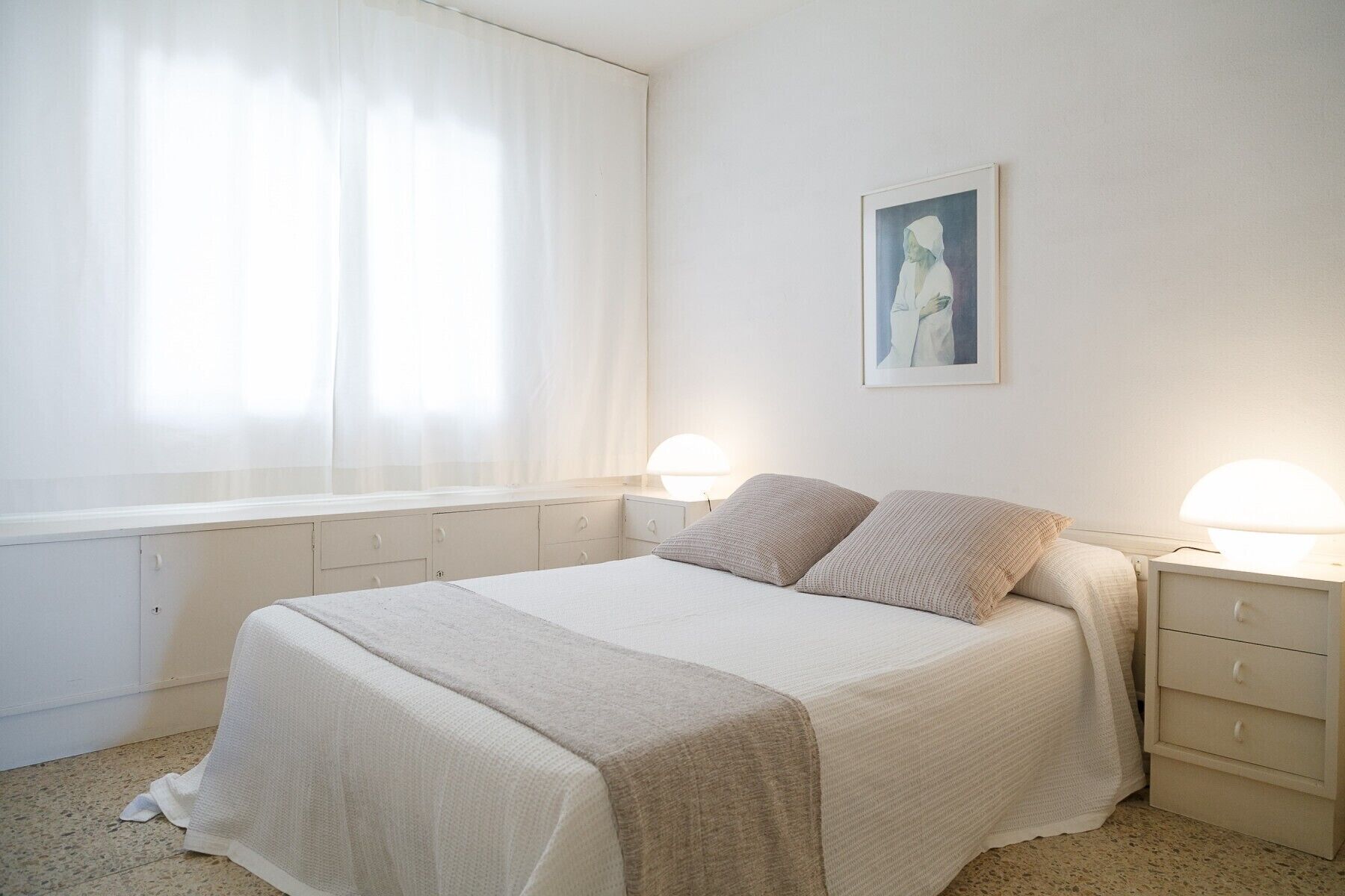 Un dormitorio con cama, dos mesitas de noche, una lámpara de noche, una ventana con cortinas translúcidas y un cuadro en la pared.