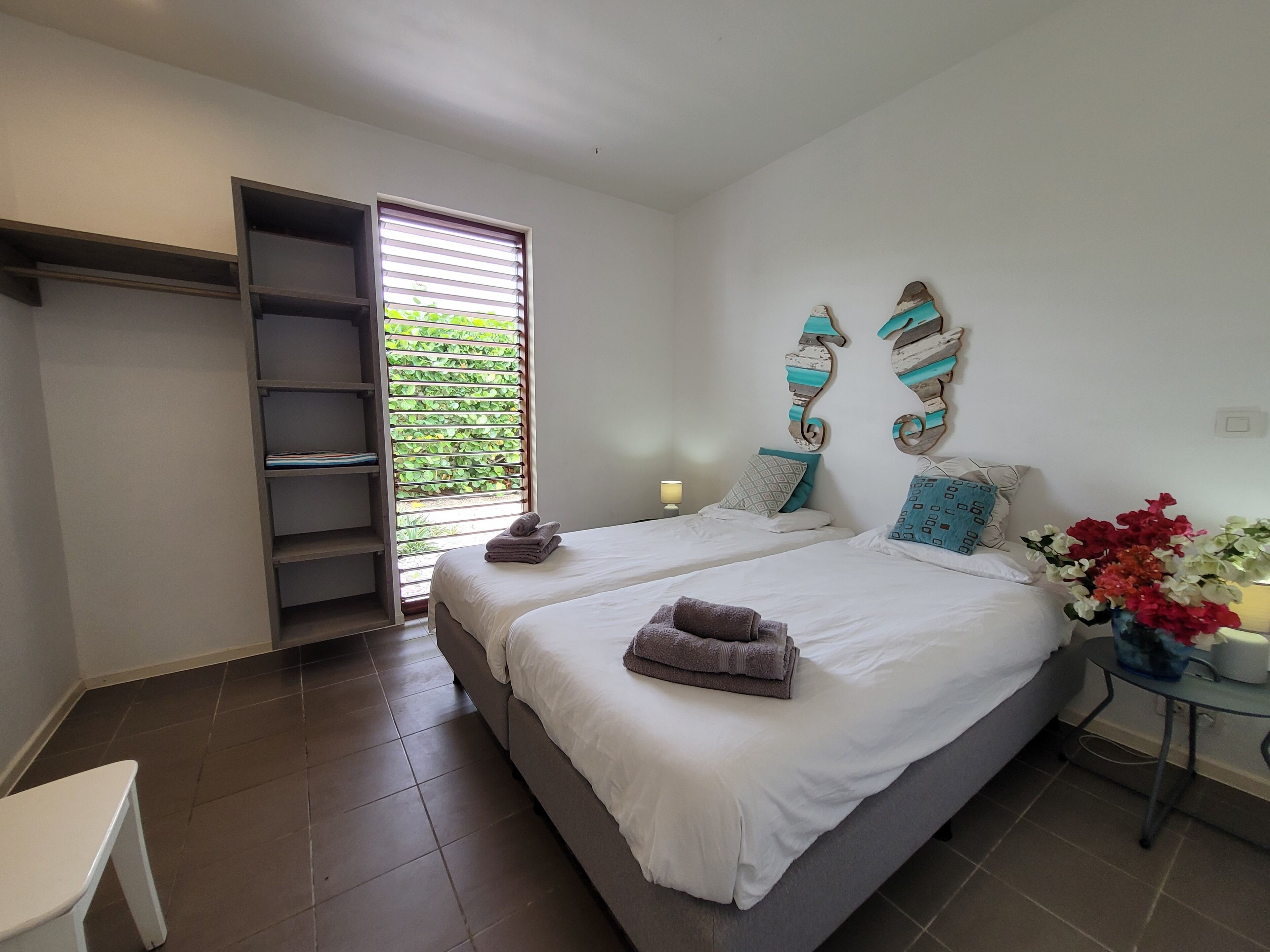 Une chambre avec deux lits, une armoire, une petite table avec une lampe et une fenêtre avec des stores.