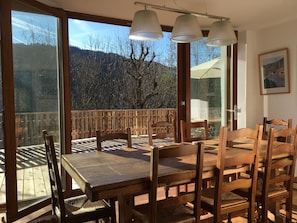 Une table à manger en bois, avec des chaises, de grandes fenêtres et une vue sur les montagnes.