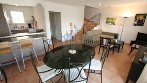 A dining area with a glass table and metal chairs, a kitchen in the background, a staircase, and a living room with a sofa and a lamp. - Villa with garden near the sea (Narbonne)