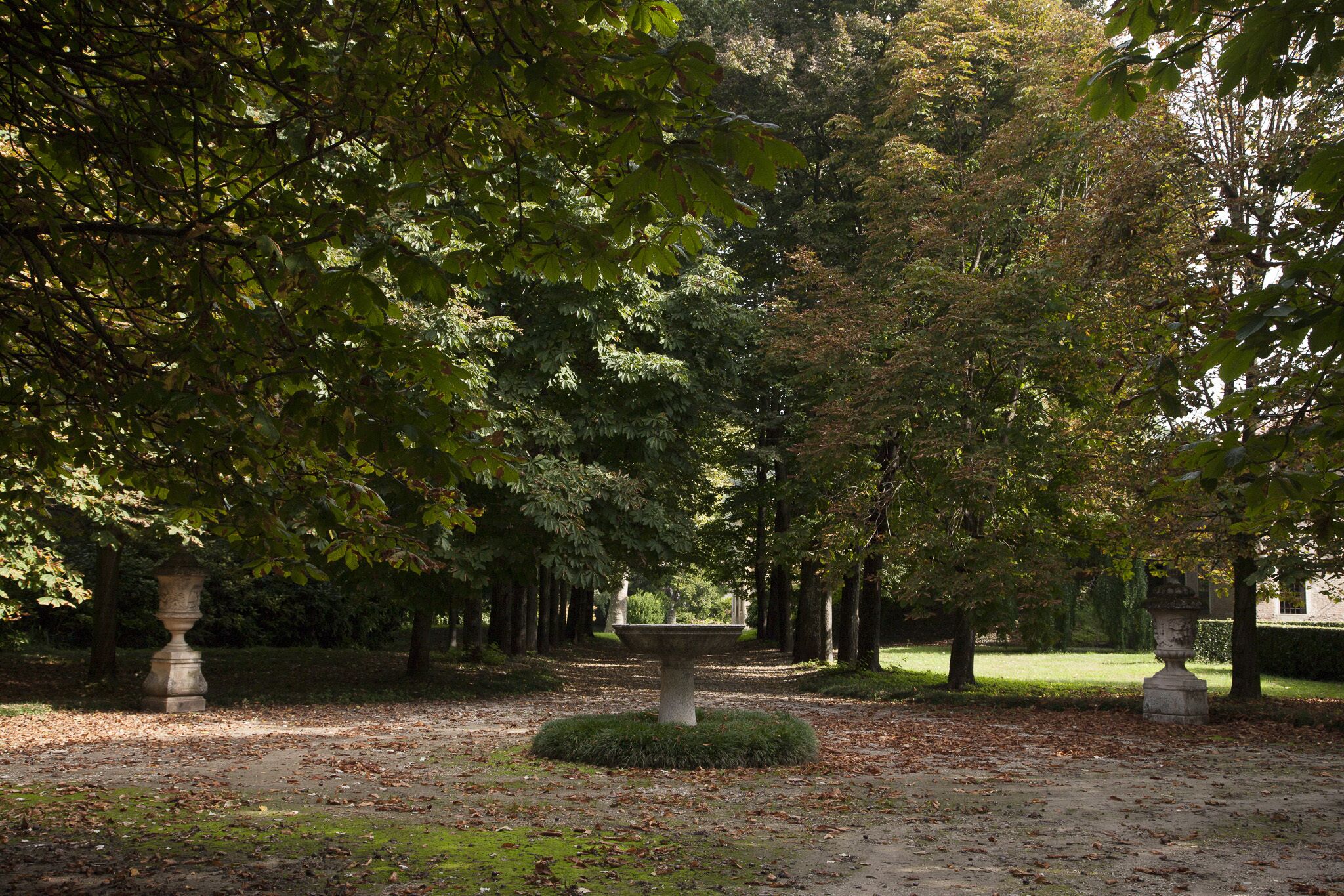 Un parc avec une fontaine centrale en pierre, entourée d’arbres et bordée d’une pelouse bien entretenue.