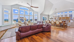 A spacious living room with a large sectional sofa, a dining area with wooden furniture, and large windows offering a view of the ocean.