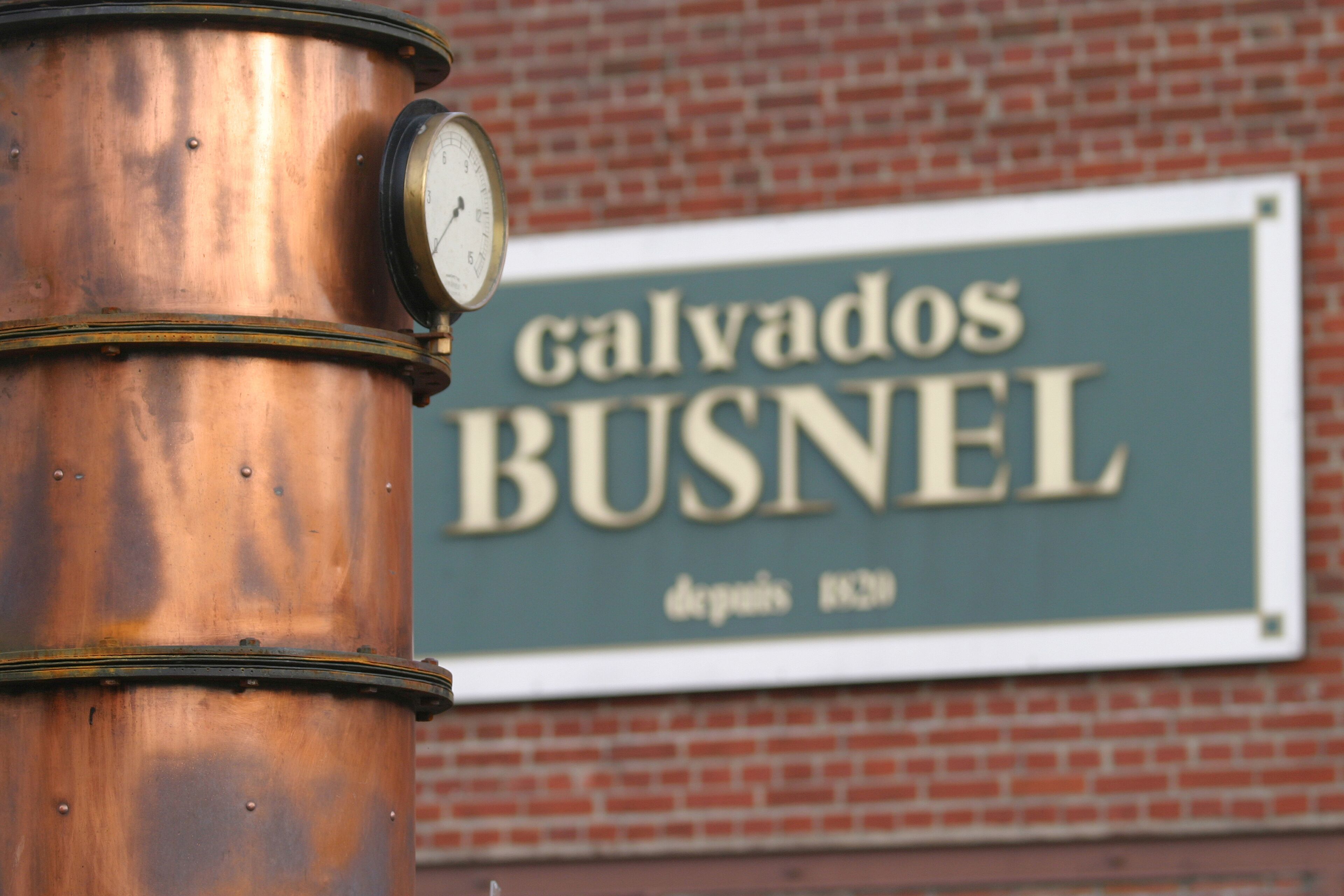 Une horloge en laiton, fixée sur un poteau, avec une enseigne en arrière-plan indiquant « Calvados BUSNEL ».