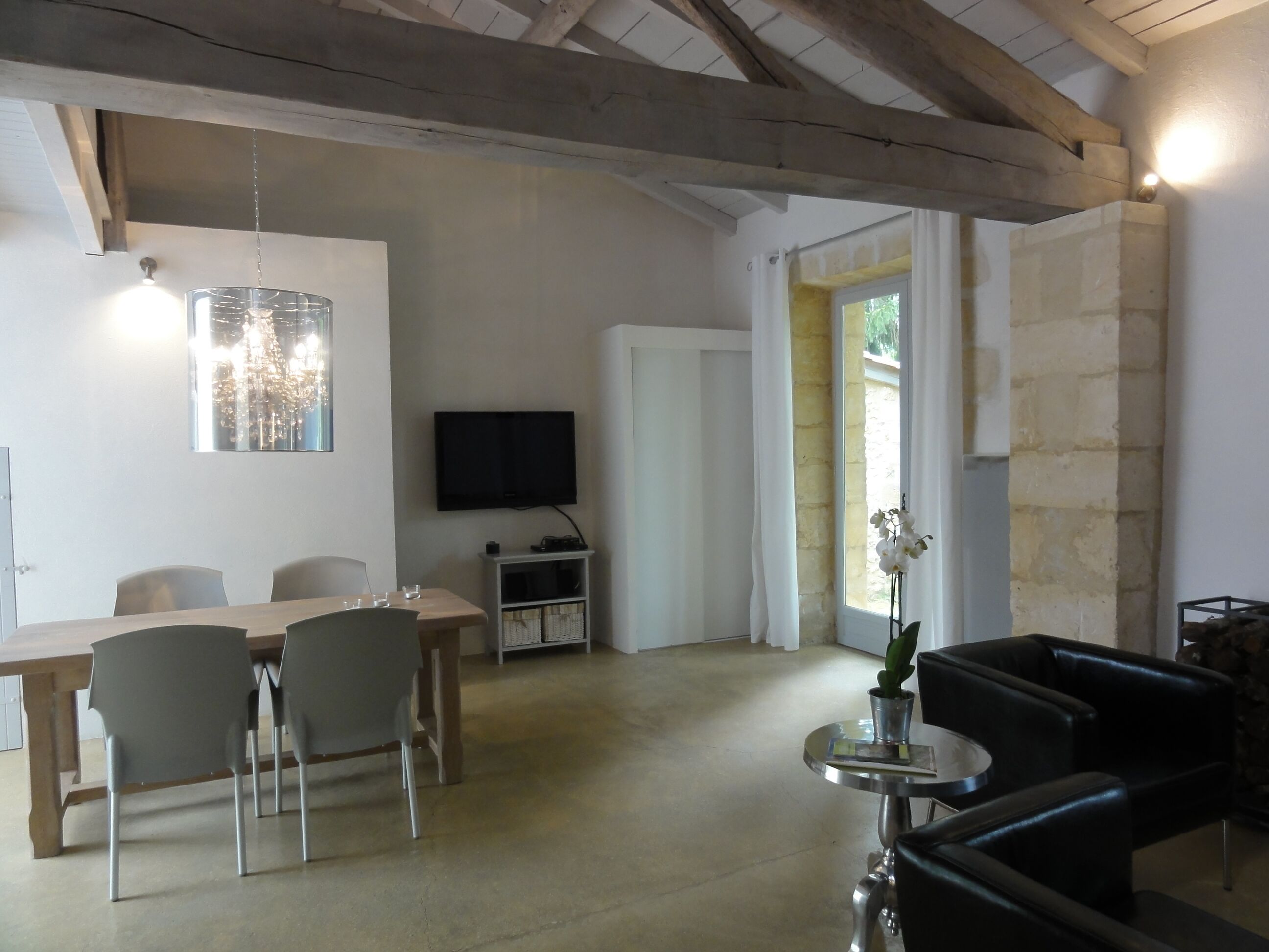 A spacious living area with a wooden dining table, chairs, a flat-screen TV, a small side table with a vase, and a black leather sofa.