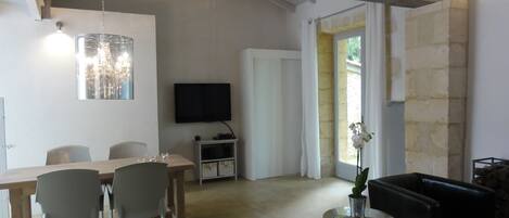 A spacious living area with a wooden dining table, chairs, a flat-screen TV, a small side table with a vase, and a black leather sofa.