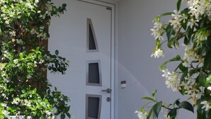 Una puerta blanca con diseño geométrico, flanqueada por macetas grandes con flores blancas.