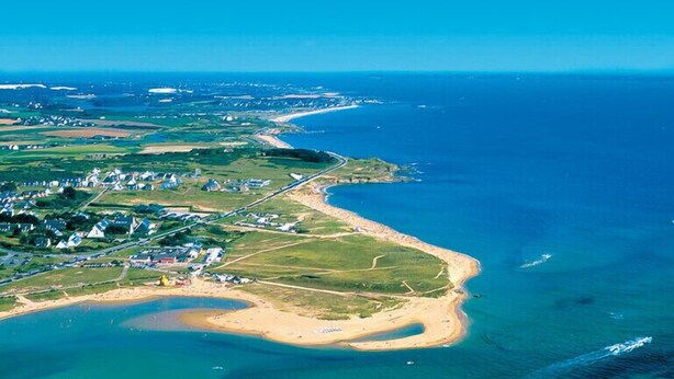Vista aérea de una zona costera con playa, un pueblo pequeño y un puerto deportivo.