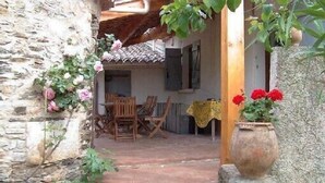 Un bâtiment rustique en pierre, avec une table et des chaises en bois, un mur de pierre orné de plantes grimpantes et un grand pot rempli de fleurs rouges.