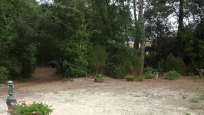 A sandy outdoor area with a wooden planter with flowers, surrounded by dense greenery and trees. - Le Verdon Sur Mer: House / Villa - Soulac / Le Verdon sur Mer (Le Verdon-sur-Mer)