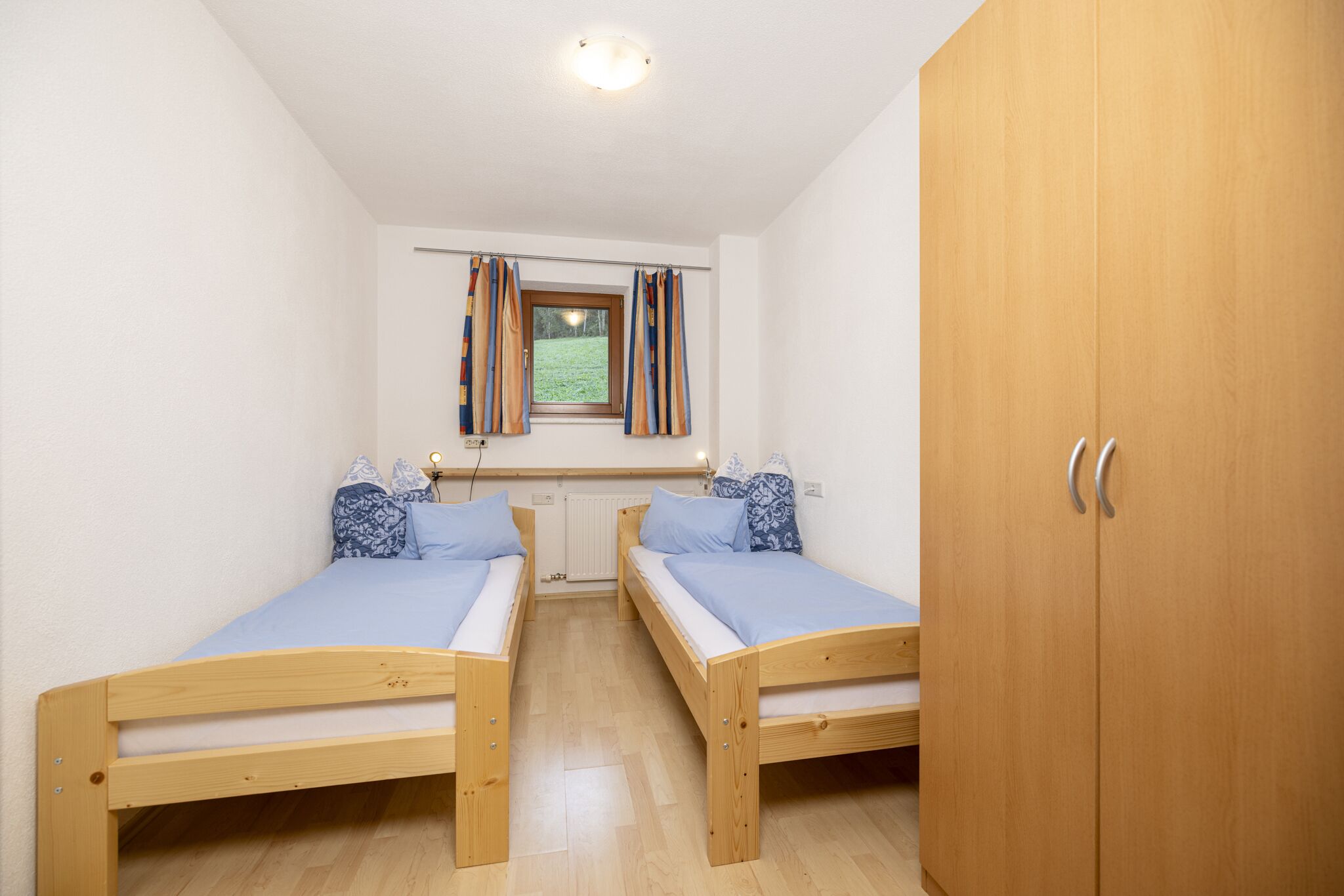 Habitació petita i ben il·luminada amb dos llits de fusta, una finestra amb cortines, un radiador i un armari.