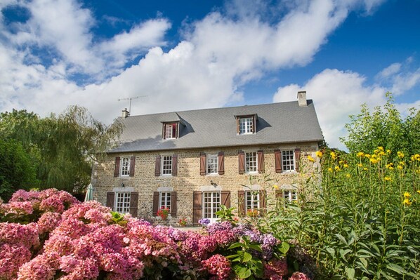 Una casa de piedra con techo gris, varias ventanas con contraventanas y un jardín con flores rosas y púrpuras.