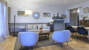 Een woonkamer met een bank, twee fauteuils, een salontafel, een eettafel met stoelen, een lamp, een spiegel en een kast met decoratieve objecten.