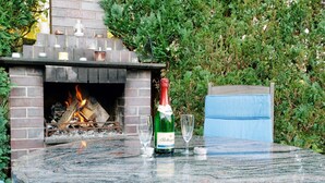 Uma lareira de pedra com fogo, uma garrafa de vinho e duas taças sobre uma mesa de mármore.