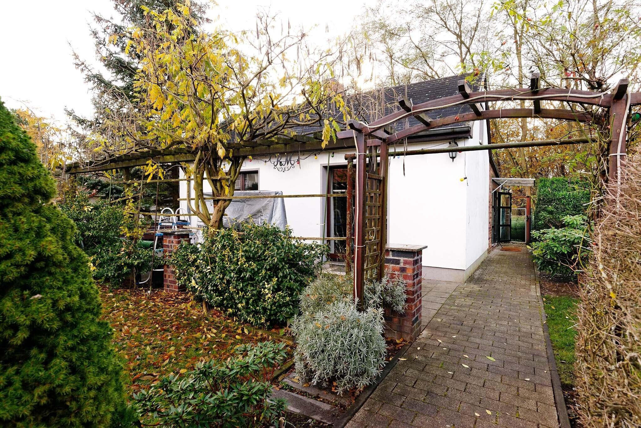 Ein weißes Gebäude mit einer Ziegelveranda, umgeben von einem Garten mit verschiedenen Sträuchern und einer Pergola.