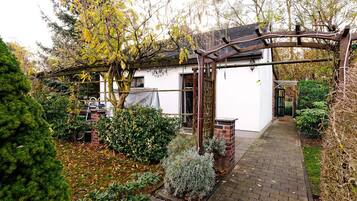 Ein weißes Gebäude mit einer Ziegelveranda, umgeben von einem Garten mit verschiedenen Sträuchern und einer Pergola.