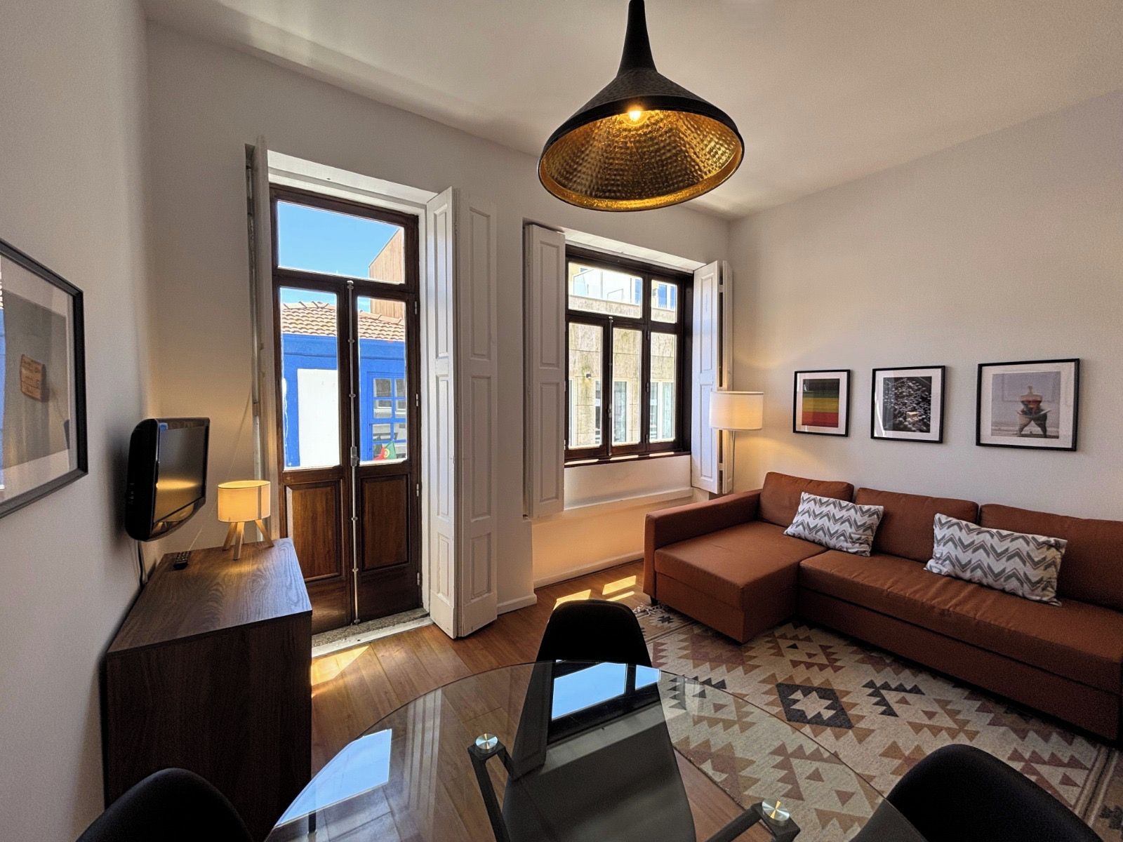 A modern living room with a glass table, brown sofa, and a hanging light fixture.
