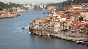 A riverside cityscape with historic buildings, a bridge, and boats docked along the waterfront.