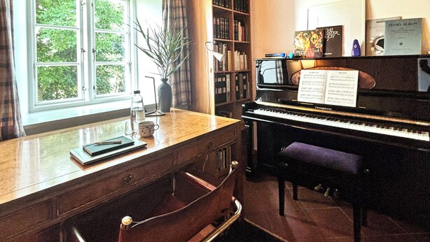 Une pièce avec un piano, un bureau avec une chaise, une bibliothèque et une fenêtre avec des rideaux.