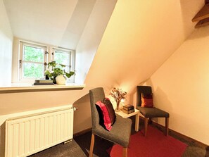 A corner room with a radiator, a window with plants, a chair, a small table, and a red carpet.