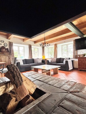 A cozy living room with a large sectional sofa, a coffee table, and a wooden log centerpiece. There are windows with curtains, a chandelier, and a fireplace.