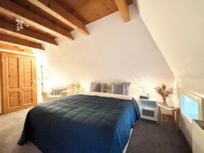 Un dormitorio con techo inclinado, una cama con cubrecama azul, una silla, una mesita de noche con lámpara y una ventana.