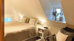A bedroom with a sloped ceiling, a bed with a striped bedspread, a bedside table with a lamp, a radiator, and a window with a plant. - Dream house under thatch with fireplace, sauna, wellness room and lots of space (Poppenbüll)