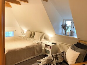 Ein Schlafzimmer mit schrägem Dach, einem Bett mit gestreifter Bettdecke, einem Nachttisch mit Lampe, einem Heizkörper und einem Fenster mit Pflanze.