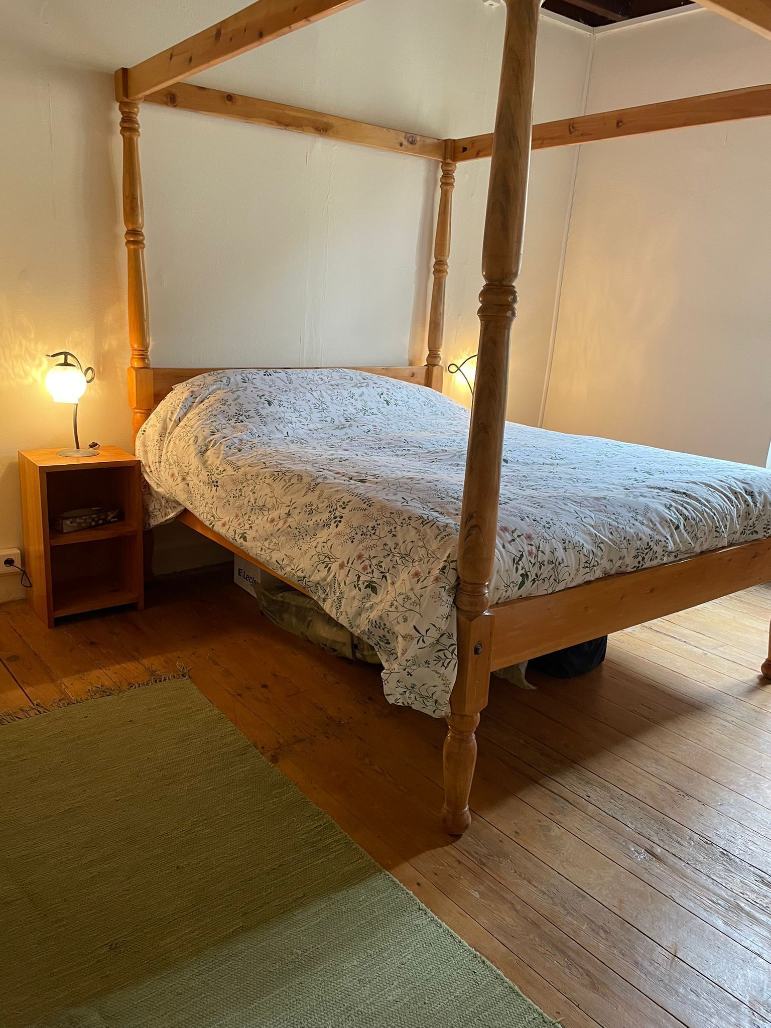 Un lit à baldaquin avec une couvre-lit à motifs floraux, une table de chevet avec une lampe et un tapis vert sur un plancher en bois.