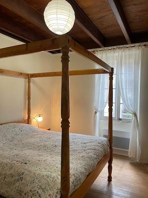 Ein Bett mit Baldachin, ausgestattet mit einem floralen Überwurf, einer Nachttischlampe und einem Fenster mit durchsichtigen Vorhängen.
