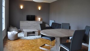 Ein modernes Wohnzimmer mit einem runden Holztisch, grauen Sofas, einem weißen Fernseher und zwei Wandleuchten.