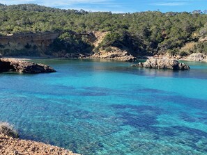 A coastal landscape with clear turquoise waters, rocky shores, and a forested hill in the background.