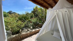 Una habitación con techo de madera, una cama con dosel blanco y vistas a un jardín con diversas plantas y un muro de piedra.