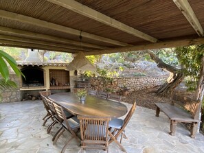 An outdoor patio with a stone floor, a wooden table with chairs, a hanging light fixture, and a stone wall with plants.