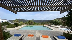 Zona de piscina con sillones reclinables y jacuzzi, rodeada de vegetación y vistas a colinas.