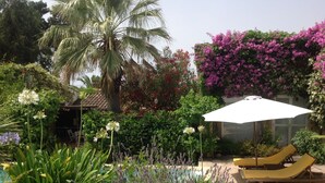 Ein Poolbereich mit Liegestühlen, einem weißen Sonnenschirm und üppiger Bepflanzung, darunter blühende Pflanzen und eine Palme.
