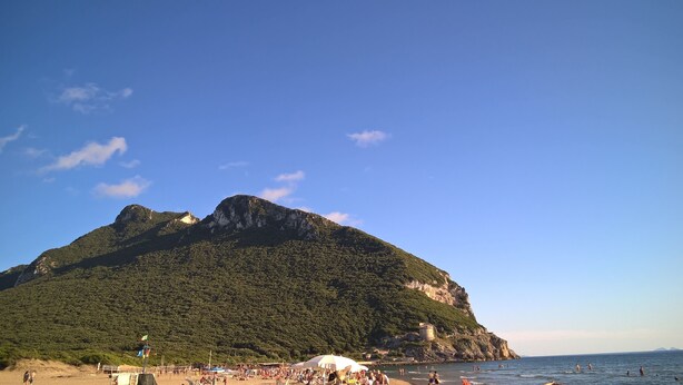 Een strand met een grote berg op de achtergrond, een heldere lucht en mensen die in het water zwemmen.
