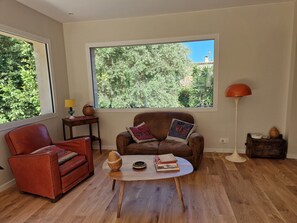A living room with a brown leather sofa, a red armchair, a wooden coffee table, and a large window with a view of trees. - Old stone house with a nice garden (Vallauris)