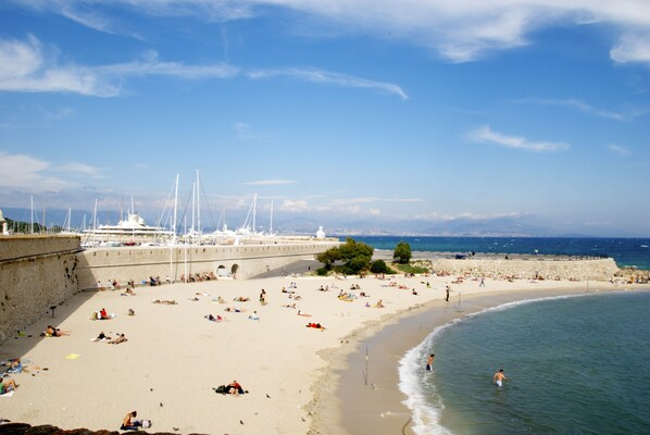 Une plage où des gens se prélassent au soleil et se baignent, une jetée avec des bateaux, et un ciel dégagé.