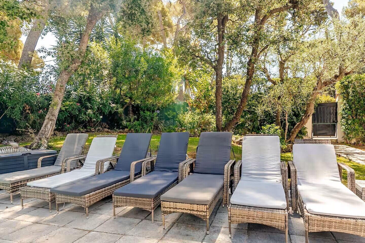 A row of wicker lounge chairs with cushions, arranged on a stone patio surrounded by trees.