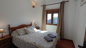 A bedroom with a wooden bed, patterned bedding, a nightstand with a lamp, a window with curtains, and a view of mountains.