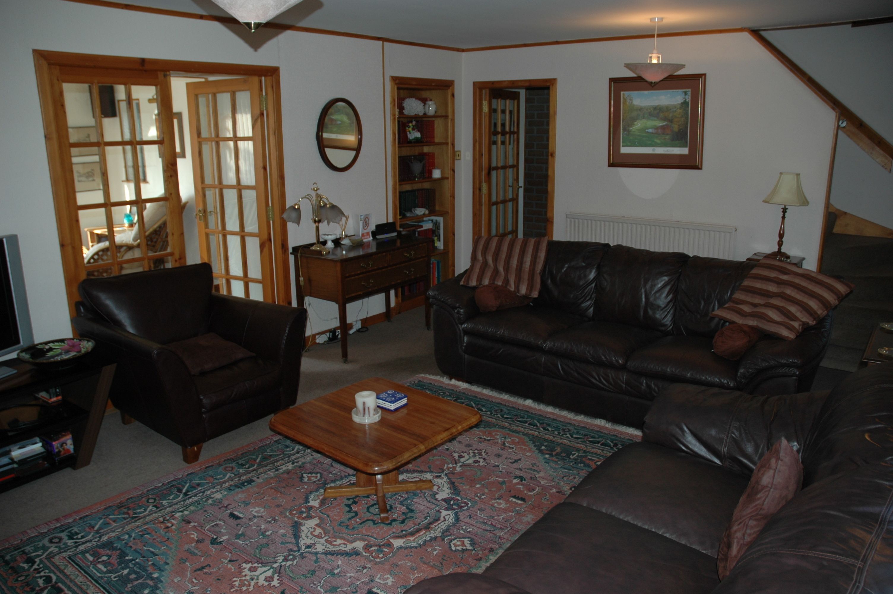 A living room with a brown leather sofa, a wooden coffee table, a patterned rug, a wooden side table, and a bookshelf.