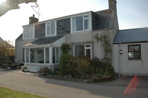 A two-story house with a porch, a chimney, and a garden with flowers. - The Old Croft (Brora)