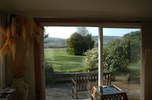 A room with a large sliding glass door leading to a view of a grassy field, a wooden bench, and a table with a book. - The Old Croft (Brora)