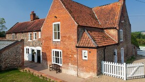 Une maison traditionnelle en briques, avec un toit à forte pente, des fondations en pierre et une clôture en bois peinte en blanc.