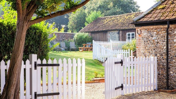 Un bâtiment rustique en pierre, avec une clôture en bois peinte en blanc, un chemin de gravier et un arbre au premier plan.