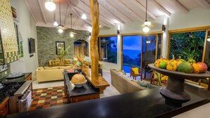 A modern kitchen with a black countertop, a wooden dining table, and a large window overlooking a scenic view. - 6BR Luxury Ocean View Villa • 3 Private Pools • Chef Included • Sky Deck (Manuel Antonio)